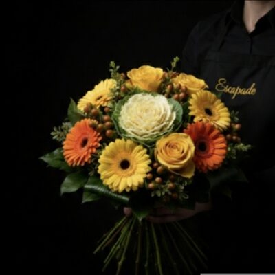 Bouquet "Cœur de Saison" ​L'Esprit de la Création ​Une composition chaleureuse et structurée qui capture l'essence même de la générosité. Ce bouquet rond serré offre un contraste élégant entre la délicatesse des fleurs nobles et le caractère rustique du chou ornemental. Ses teintes vitaminées apporteront une lumière instantanée à votre intérieur ou feront le bonheur d'un proche pour toute occasion festive. ​Détails de la Composition ​Chaque bouquet est réalisé avec soin par nos artisans : ​Pièce Maîtresse : Un chou d'ornement majestueux placé au cœur de la création pour une structure unique. ​Fleurs Majeures : Une sélection de roses jaunes lumineuses et de germinis éclatants déclinés en jaune et orange. ​Finitions : Quelques baies de saison pour apporter relief et texture à l'ensemble. ​Écrin Végétal : Le bouquet est souligné par un feuillage de collerette dense, garantissant une finition parfaite et une excellente tenue.