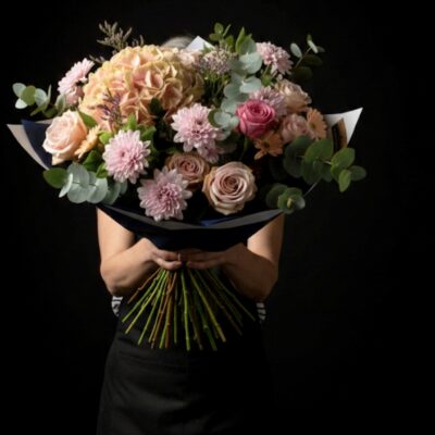 Bouquet "Nuances de Rose" ​L'Esprit de la Création ​Une composition majestueuse qui célèbre la tendresse et la féminité dans toute leur splendeur. Ce bouquet volumineux joue sur un dégradé de roses délicats, offrant une texture riche et une profondeur visuelle captivante. C'est une pièce d'exception, idéale pour exprimer de grands sentiments ou pour apporter une touche d'élégance poétique à un intérieur. ​Détails de la Composition ​Chaque création est une pièce unique, travaillée avec une générosité florale absolue : ​La Pièce Maîtresse : Un hortensia rose somptueux aux inflorescences denses, apportant un volume spectaculaire et une base texturée. ​Le Cœur de Roses : Une sélection de roses  dans différentes teintes (poudré, vieux rose et rose tendre) pour créer un relief harmonieux. ​Touches Délicates : Des fleurs de saison coordonnées pour parfaire la rondeur et la densité du bouquet. ​Écrin Végétal : Un travail sur le feuillage d'eucalyptus aux nuances argentées qui souligne la fraîcheur des fleurs tout en diffusant un parfum apaisant. ​Finition : Un emballage bleu nuit raffiné qui crée un contraste élégant et met en lumière l'éclat des nuances rosées.
